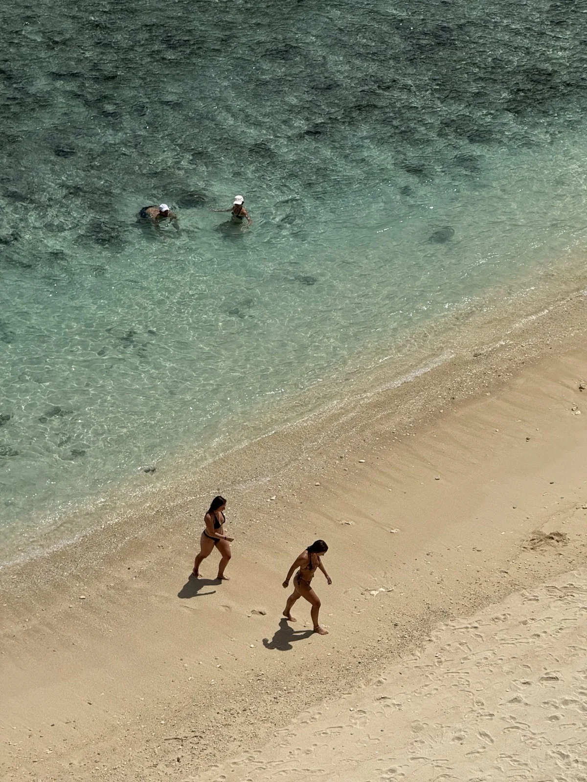 Aerial view of two travellers walking a pale sand beach in Bali beside clear turquoise shallows, with snorkellers in the water